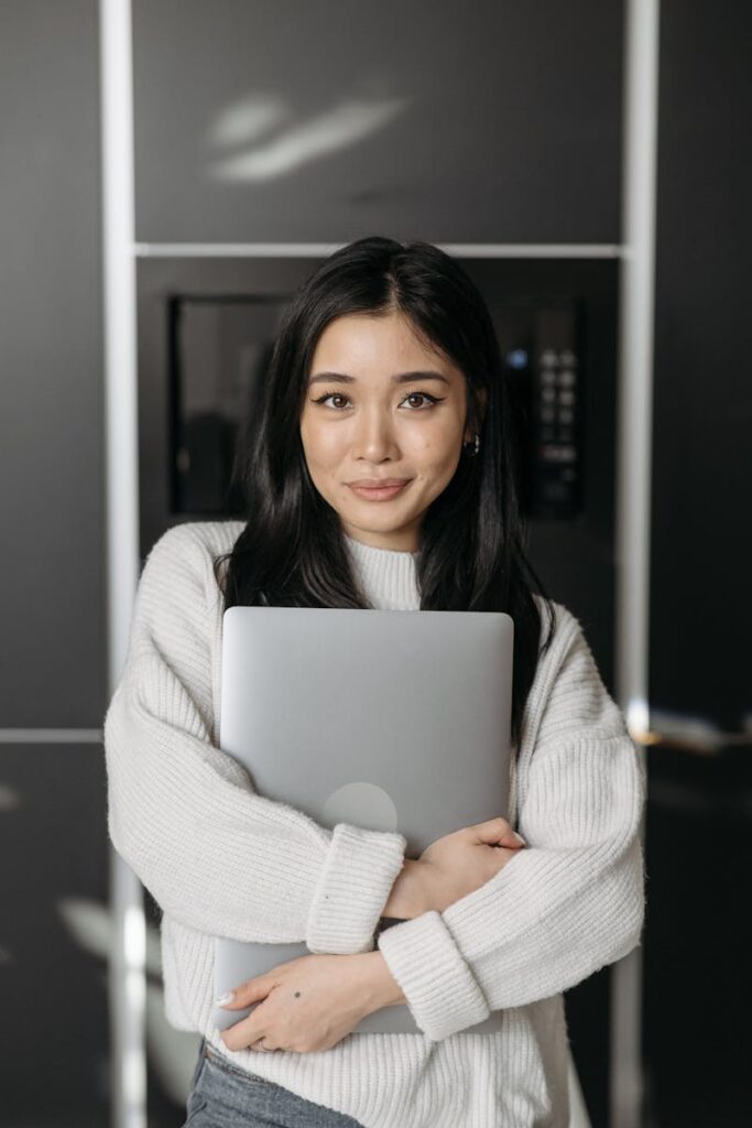 Asian woman smiling and embracing laptop inside a modern home setting.